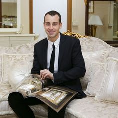 a man in a suit sitting on a couch holding a book and smiling at the camera