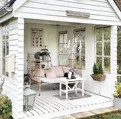 a small white house with a couch and table in the front yard, surrounded by greenery