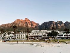 people are walking on the beach in front of some buildings and palm trees with mountains in the background