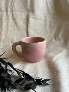 a pink coffee cup sitting on top of a white table cloth next to a plant