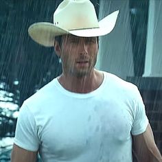 a man in a white shirt and cowboy hat standing in the rain with his hand on his hip