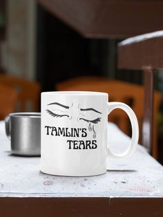 a white coffee mug with the words tamlin's tears on it next to a metal container