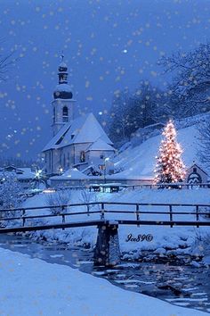 a christmas tree is lit in front of a snowy church and bridge with snow falling on the ground