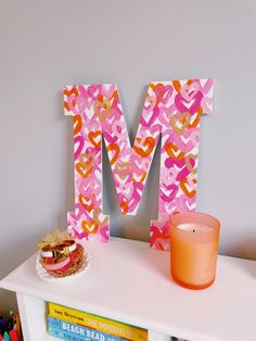 the letter m is made out of pink and orange paper hearts on a shelf next to a candle