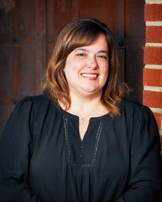 a woman in black shirt standing next to a brick wall and smiling at the camera