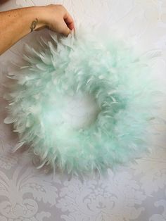 a hand holding a green feather wreath on top of a white wallpapered surface