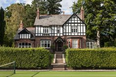 a tennis court in front of a large house with hedges on each side and a tennis net at the end