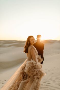 a man and woman standing in the sand at sunset