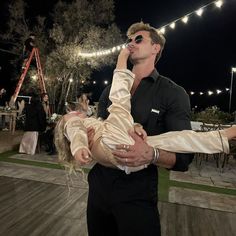 a man holding a woman in his arms at an outdoor event with string lights on the background