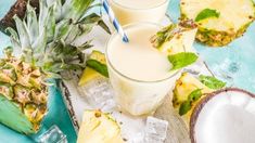 three glasses filled with pineapple milk next to sliced pineapples and ice cubes