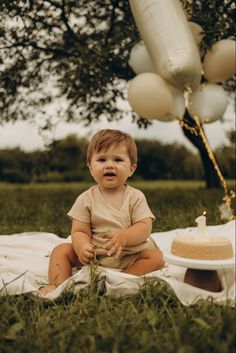 a baby sitting in the grass next to a cake with candles on it and balloons