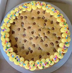 a cookie cake with yellow frosting and chocolate chips on the top is in a box