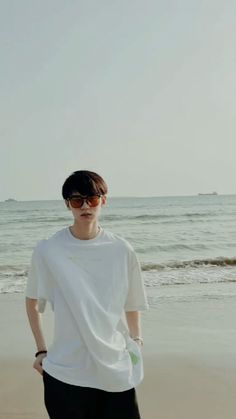 a young man standing on top of a sandy beach next to the ocean with his hands in his pockets