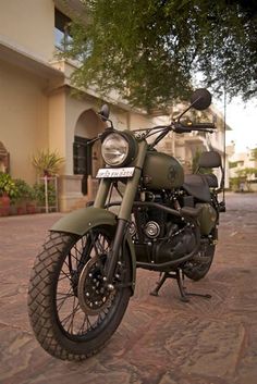 a green motorcycle parked in front of a building on a brick sidewalk with trees and bushes