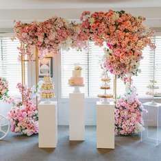 a table topped with lots of pink flowers next to tall white pedestals filled with cakes