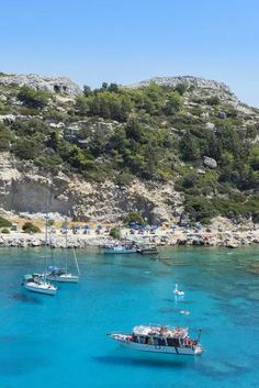 several boats floating in the blue water next to a rocky shore with trees on it