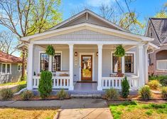 a small white house with porches and plants on the front door is surrounded by trees