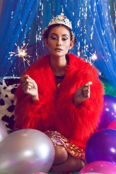 a woman in a red fur coat holding sparklers while sitting next to balloons and streamers