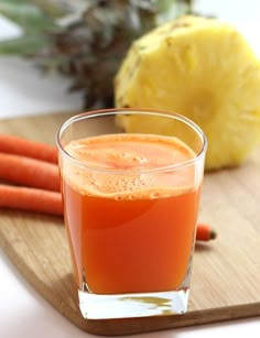 a glass filled with orange juice next to some carrots and pineapple on a cutting board