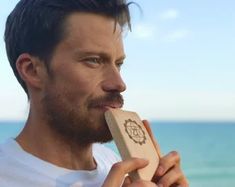 a man is brushing his teeth by the ocean with an object in his hand and looking off into the distance