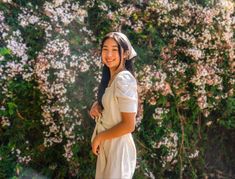 a woman in white dress standing next to a bush with lots of flowers on it