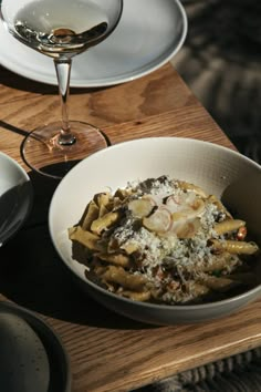 a white bowl filled with pasta on top of a wooden table next to a glass of wine