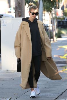 a woman walking down the street wearing a tan coat and black top with white sneakers