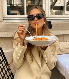 a woman in sunglasses is eating spaghetti from a white bowl with a fork and spoon