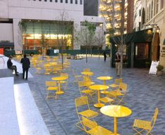 several yellow chairs and tables in an open area with people walking around the building behind them