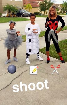 three children in costumes standing on the sidewalk with scissors and paper cutouts that spell out shoot