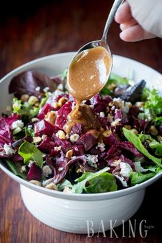 a person pouring dressing onto a salad in a white bowl on a wooden table with lettuce and other vegetables