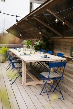 an outdoor dining table with blue chairs and lights strung over it on a wooden deck