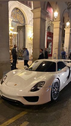 a white sports car parked on the street in front of a building with people standing around it