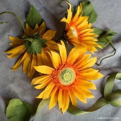 three sunflowers with green stems and leaves on a table cloth next to ribbons