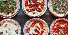 six pizzas with different toppings are arranged in rows on a wooden table top