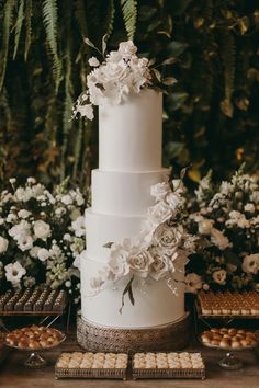 a white wedding cake with flowers on top and cookies in the foreground, surrounded by greenery