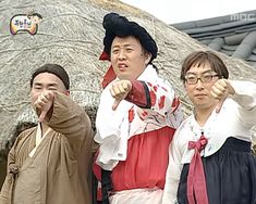 three people standing next to each other in front of a thatched roof with one pointing at the camera