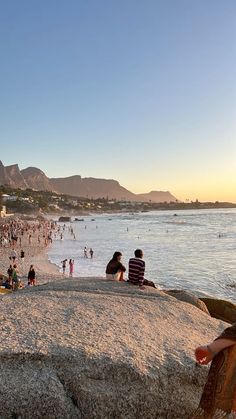 many people are on the beach and in the water at sunset or sunrise, with mountains in the background