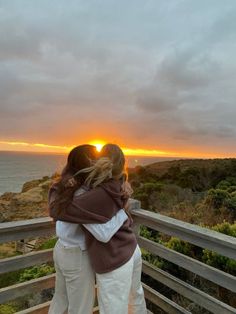 two people embracing each other while standing on a wooden deck overlooking the ocean at sunset