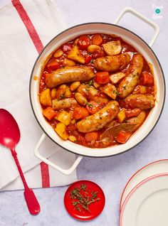 a pan filled with sausages and potatoes on top of a table next to a red spoon