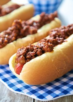 three chili cheese dogs on a blue and white checkered tablecloth with other hotdogs in the background