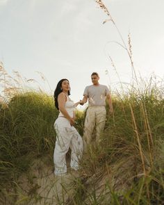 a man and woman standing on top of a sandy beach next to tall green grass