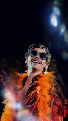 a man with sunglasses and feathers on his shoulders, singing into a microphone in front of bright lights