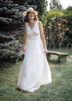 a woman in a white dress and hat is walking through the grass near a bench