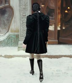 a woman in black coat and boots walking on snow covered ground next to large building