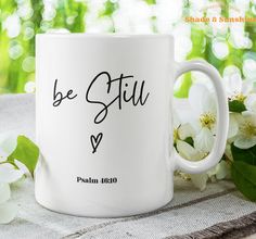 a white coffee mug with the words be still on it next to flowers and leaves