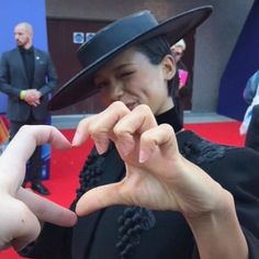 a woman in a hat making a heart sign with her hands while standing on a red carpet