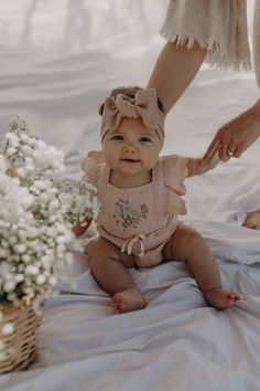 a baby sitting on a bed next to a basket with flowers and a woman holding a flower pot