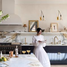 a woman in a white dress standing at a kitchen counter with wine glasses on it