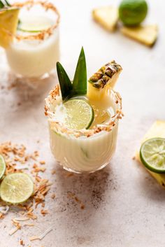 two glasses filled with drinks and garnished with limes on a table next to sliced pineapple slices